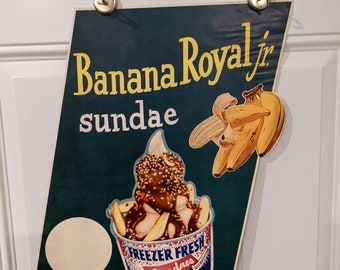 Può includere: Una pubblicità vintage per i sundae Banana Royal Jr. La pubblicità presenta un'illustrazione colorata di un sundae alla banana con salsa al cioccolato, panna montata e codette. Il testo "Freezer Fresh Sundaes" è stampato sul lato del bicchiere del sundae. La pubblicità presenta anche un grappolo di banane gialle.