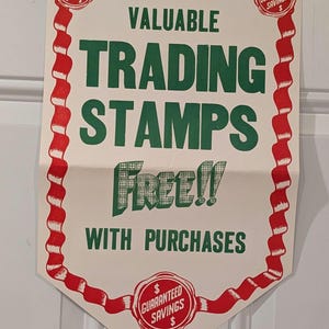 Puede incluir: Un cartel blanco con bordes de cinta roja y blanca. El cartel anuncia "VALUABLE TRADING STAMPS Free!! WITH PURCHASES". Tres sellos rojos y blancos con el texto "GUARANTEED SAVINGS" están en el cartel.