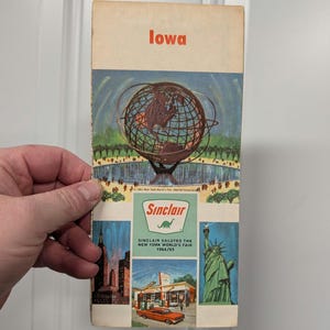 Könnte beinhalten: Vintage Iowa Reisebroschüre mit einer Globusillustration und dem Text "Iowa" in Rot. Die Broschüre enthält Bilder der New Yorker Weltausstellung und Sinclair Tankstellen. Das Sinclair-Logo zeigt einen grünen Dinosaurier.