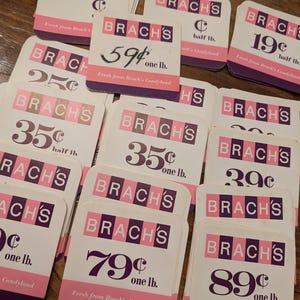Puede incluir: Una pila de etiquetas de precios de caramelos Brach's vintage. Las etiquetas son blancas con letras moradas y rosas. Los precios van desde 19 centavos hasta 89 centavos por libra. Las etiquetas dicen "Fresh from Brach's Candyland".