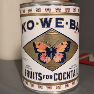May include: A vintage metal can of Ko-We-Ba brand fruits for cocktail. The can is white with a gold and blue label featuring a butterfly and the text "Fruits for Cocktail".