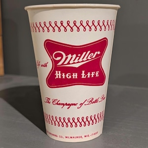 Puede incluir: Un vaso de papel vintage de Miller High Life. El vaso es blanco con detalles en rojo, incluido un patrón de remolino repetido y el logotipo de Miller High Life en un escudo rojo. El texto en el vaso dice "The Champagne of Bottle Beer."