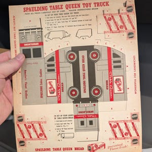May include: A vintage cardboard toy truck die-cut from a box of Spaulding Table Queen Bread. The truck is red, white, and gray with black wheels. The box has a red and white design with the words "Spaulding Table Queen Bread" printed on it. The instructions for assembling the truck are printed on the back of the box.