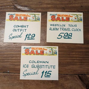 Könnte beinhalten: Drei Vintage-Verkaufsschilder mit einer Cartoon-Illustration von Personen, die einen Urlaub genießen. Die Schilder werben für ein Kampfoutfit für 129 $, eine Westclox Tour Alarm-Reiseuhr für 5,98 $ und einen Coleman-Eissubstitut für 1,15 $.