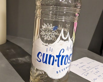 May include: A vintage glass bottle with a white label that says "sun-frost beverage" in blue lettering. The bottle is clear and has a textured surface. The label also features a sun with a smiling face and a mountain range.