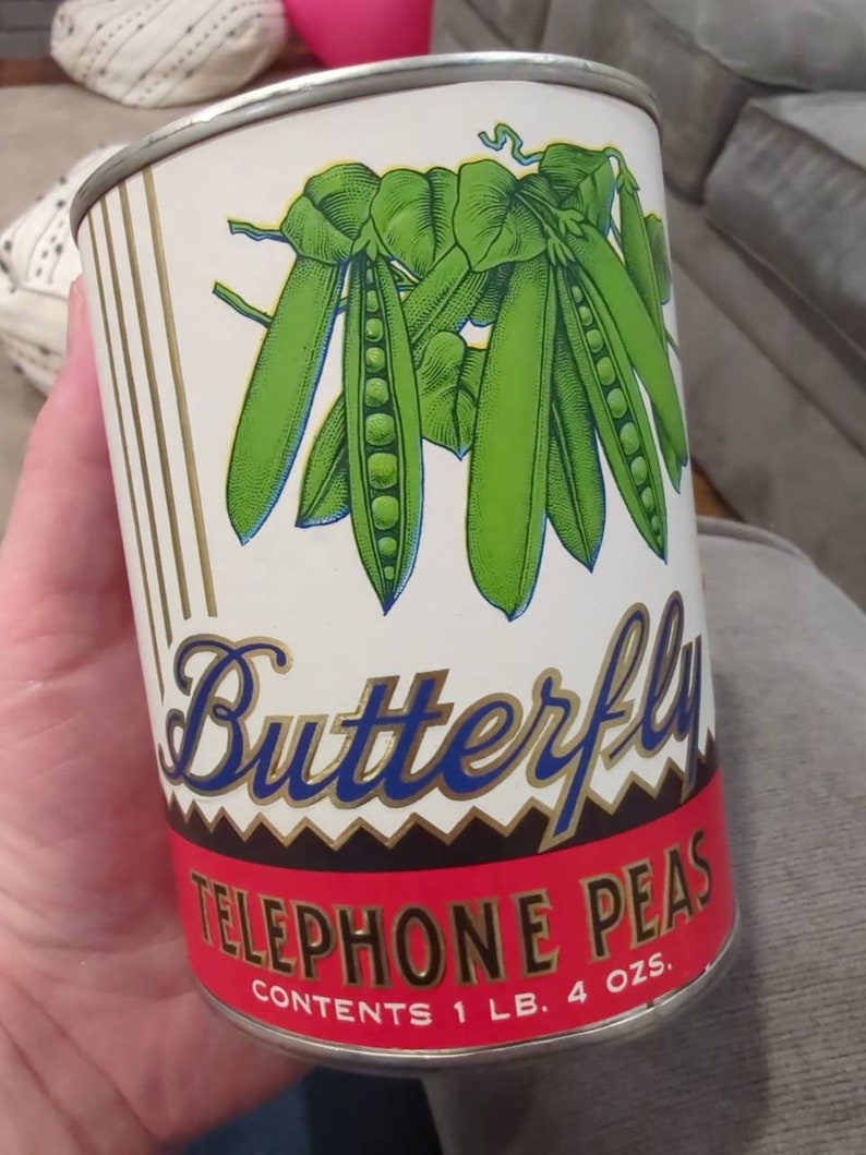 May include: A vintage can of Butterfly Telephone Peas. The can is white with a red, black, and gold label. The label features a drawing of green peas in pods with leaves. The text on the label reads "Butterfly Telephone Peas" and "Contents 1 lb. 4 oz."
