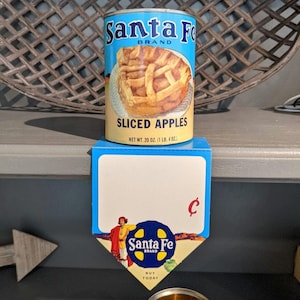 May include: A vintage can of Santa Fe brand sliced apples. The can is blue and white with a picture of a sliced apple pie on the front. The can is sitting on a shelf with a price tag that says "C".