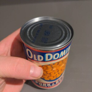 May include: A can of Old Dominion Pork & Beans with a blue and red label. The can is made of metal and has a silver lid. The label says "Old Dominion" in blue letters and "Since 1885" in smaller blue letters. The can is partially filled with beans.