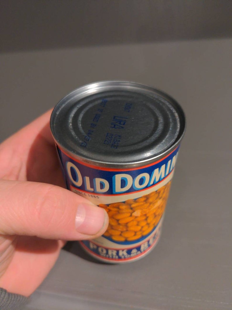 May include: A can of Old Dominion Pork & Beans with a blue and red label. The can is made of metal and has a silver lid. The label says "Old Dominion" in blue letters and "Since 1885" in smaller blue letters. The can is partially filled with beans.