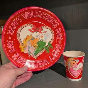 Puede incluir: Un juego de plato y vaso de papel rojo con temática de San Valentín. El plato presenta a Miss Piggy y Kermit la Rana con el texto "Happy Valentine's Day". La taza tiene un diseño similar con Miss Piggy y el texto "Happy Valentine's Day!"