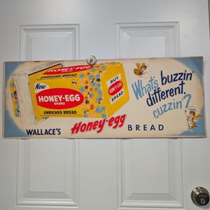 May include: A vintage advertisement for Honey-Egg bread. The ad features a yellow box of bread with the text "New Honey-Egg Brand Enriched Bread" and "Buy Honey-Egg Bread". The ad also features a cartoon bee and the text "What's buzzin' different, cuzzin?" and "Wallace's Honey-Egg Bread".