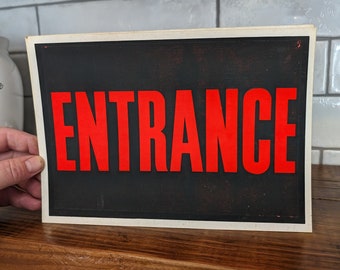 Op de afbeelding: Een zwart-wit bord met het woord "ENTRANCE" in rode letters.