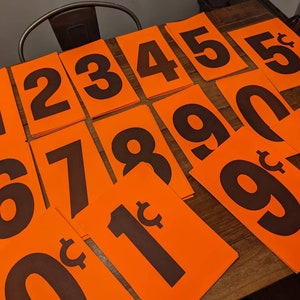 May include: A set of orange price tags with black numbers and cent signs. The numbers range from 0 to 9 and include a 1 cent tag.