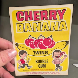 May include: A yellow and red wrapper for Cherry Banana Twins Bubble Gum. The wrapper features a banana with red cherries on top and two cartoon children. The text on the wrapper reads "Cherry Banana Twins Bubble Gum".