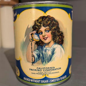 May include: Vintage tin can with a blue and yellow label featuring a young girl holding a pocket watch to her ear. The label reads "California Packing Corporation, Main Office, San Francisco, California, USA. Packed without sugar - Sweeten to taste."