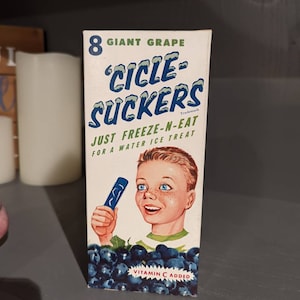 May include: A box of 8 giant grape 'Cicle-Suckers' water ice treats. The box features a cartoon boy holding a blue popsicle and the text 'Just Freeze-N-Eat' and 'Vitamin C Added'.