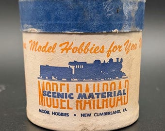 May include: A cylindrical cardboard container with a blue lid and cream-colored body. The label features a blue steam locomotive graphic, orange text reading Model Hobbies for You, and black text stating Model Railroad Scenic Material.