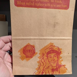 May include: A brown paper bag with a red and white logo that says "SHELL" and a red and white graphic of a Shell gas station attendant. The bag has the text "USE this LITTERBAG for trash. When soiled replace with new one from" printed on the front.