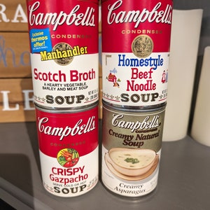 Könnte beinhalten: Vier rote und weiße Campbell's Suppendosen. Die Dosen sind übereinander gestapelt. Die oberste Dose ist Cremige Natur Suppe, die zweite Dose ist Knuspriger Gazpacho, die dritte Dose ist Schottischer Eintopf und die unterste Dose ist Hausgemachte Rindfleischnudelsuppe.
