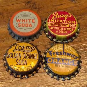 Puede incluir: Cuatro tapas de botellas de refrescos vintage. Las tapas son de color naranja, rojo, amarillo y azul. Las tapas tienen texto que dice "WHITE SODA", "Barg's IMITATION STRAWBERRY SODA", "Try-me GOLDEN ORANGE SODA" y "Freshway ORANGE BEVERAGE".