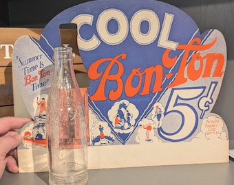 May include: A vintage cardboard display for Bon Ton soda. The display is shaped like a heart and features a blue and white background with the words "Cool Bon Ton 5¢" in red and white. The display also includes a clear glass bottle of Bon Ton soda. The text "Summer Time Is Bon Ton Time" is visible on the left side of the display.