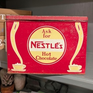 May include: Vintage red Nestlé's Hot Chocolate tin. The rectangular tin features a central yellow circle with the Nestlé's logo and the words "Ask for Hot Chocolate." Yellow streams flow from the top to teacups at the bottom.