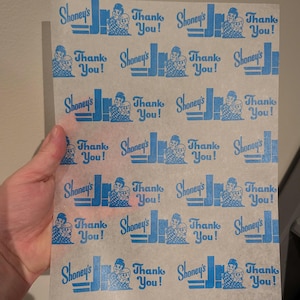 May include: A sheet of thin, translucent paper printed with a repeating pattern in blue. The design includes the text "Shoney's" and "Thank You!" along with a cartoon image of two people. The paper is held in a hand.