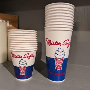 May include: Two stacks of white paper cups with a red, white, and blue design. The cups feature the words "Mister Softee" in red script, a soft-serve ice cream cone graphic, and the phrase "The Very Best."