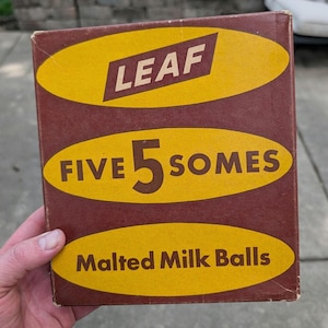 Op de afbeelding: Een vintage doos LEAF FIVE SOMES Malted Milk Balls. De doos is bruin met gele ovale accenten en tekst. Het LEAF-logo staat in witte letters in een gele ruit. De tekst "FIVE 5 SOMES" staat in een gele ovaal. "Malted Milk Balls" staat in een gele ovaal.