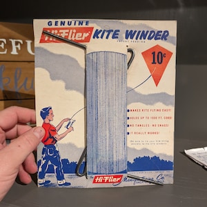 May include: A vintage Hi-Flier kite winder, a metal device for winding kite string, is shown on a blue and white illustrated cardboard package. The package features a boy flying a kite and the text "Genuine Hi-Flier Kite Winder, Patent Pending, 10c".