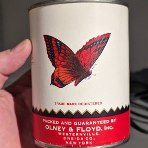 May include: A white metal can with a red and black butterfly design. The can has black text that reads "TRADE MARK REGISTERED", "PACKED AND GUARANTEED BY OLNEY & FLOYD, INC.", "WESTERNVILLE, ONEIDA CO., NEW YORK".
