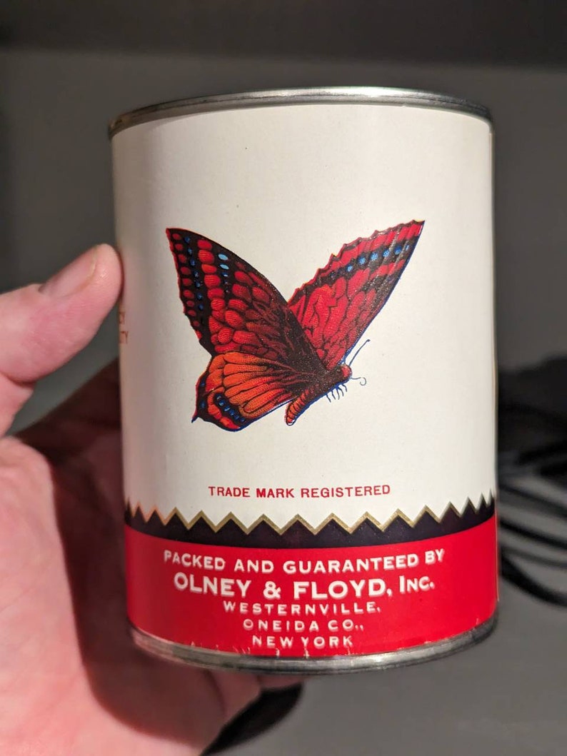 May include: A white metal can with a red and black butterfly design. The can has black text that reads "TRADE MARK REGISTERED", "PACKED AND GUARANTEED BY OLNEY & FLOYD, INC.", "WESTERNVILLE, ONEIDA CO., NEW YORK".