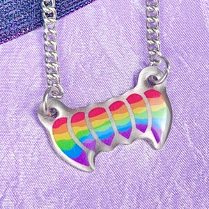 May include: A silver-toned necklace with a rainbow-colored pendant. The pendant has a curved shape with a series of rounded, tooth-like designs, each displaying a different color of the rainbow. The chain is a simple curb style, and the background is a soft purple fabric.