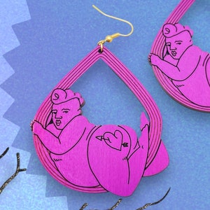 May include: A pair of pink wooden earrings shaped like a person in a yoga pose. The earrings are dangling and have a gold hook.