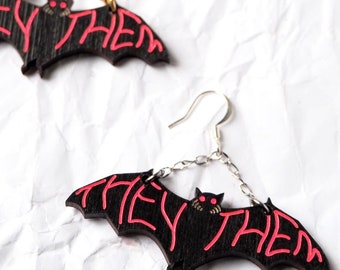 May include: A pair of black bat earrings with gold chains and red "they/them" text. The bats have red eyes and are hanging from a white surface.