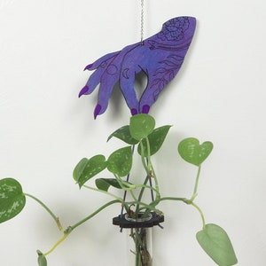 May include: A purple wooden hand with intricate designs hangs from a silver chain and holds a glass test tube filled with a plant. The plant has green leaves and is growing in the test tube.