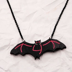 May include: A black bat pendant necklace with red detailing. The bat has red eyes and a red mouth. The necklace is on a white crumpled background.