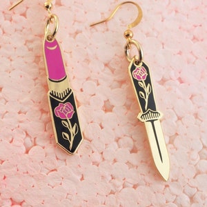May include: A pair of gold earrings with a pink lipstick and a black knife design. Each earring features a pink rose with a green stem.