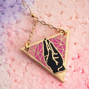 May include: Gold triangle earring with a pink and black enamel design featuring a hand gesture. The earring has a chain and a hook closure.