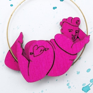 May include: A pink wooden earring featuring a figure of a person in a relaxed pose. The earring is suspended from a gold hoop.