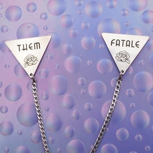 May include: A silver chain necklace with two triangular pendants. The pendants have the words "THEM" and "FATALE" engraved on them, along with a small rose design.