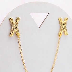 May include: A pair of gold-tone metal clasps connected by a gold chain. The clasps have a decorative, geometric design.
