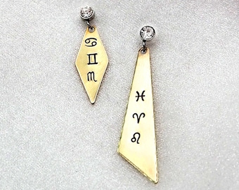 May include: A pair of gold-toned earrings with a diamond-shaped charm and a long, triangular charm. The diamond-shaped charm has the zodiac symbols for Gemini, Pisces, and Virgo engraved on it. The triangular charm has the zodiac symbols for Pisces, Aries, and Leo engraved on it.