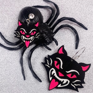 May include: A pair of black cat earrings with pink details. The earrings are shaped like a cat's face with a large, toothy grin and pink eyes. One earring is attached to a black spider charm.