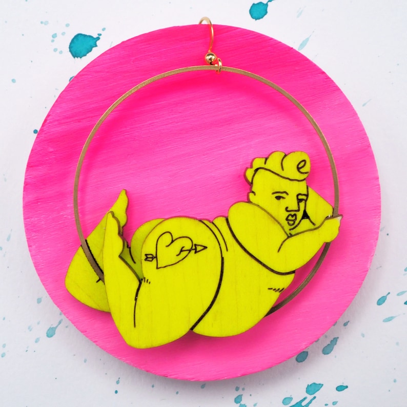May include: A pair of yellow earrings shaped like a person with a heart and arrow tattoo on their backside. The earrings are hanging from a gold hoop on a pink background.