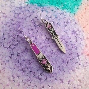 May include: A pair of silver earrings with a rose and knife design. One earring features a lipstick-shaped charm with a rose and stem, while the other earring features a knife-shaped charm with a rose and stem.