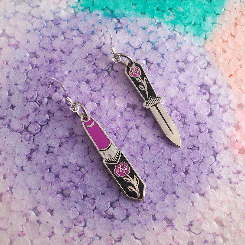 May include: A pair of silver earrings with a rose and knife design. One earring features a lipstick-shaped charm with a rose and stem, while the other earring features a knife-shaped charm with a rose and stem.
