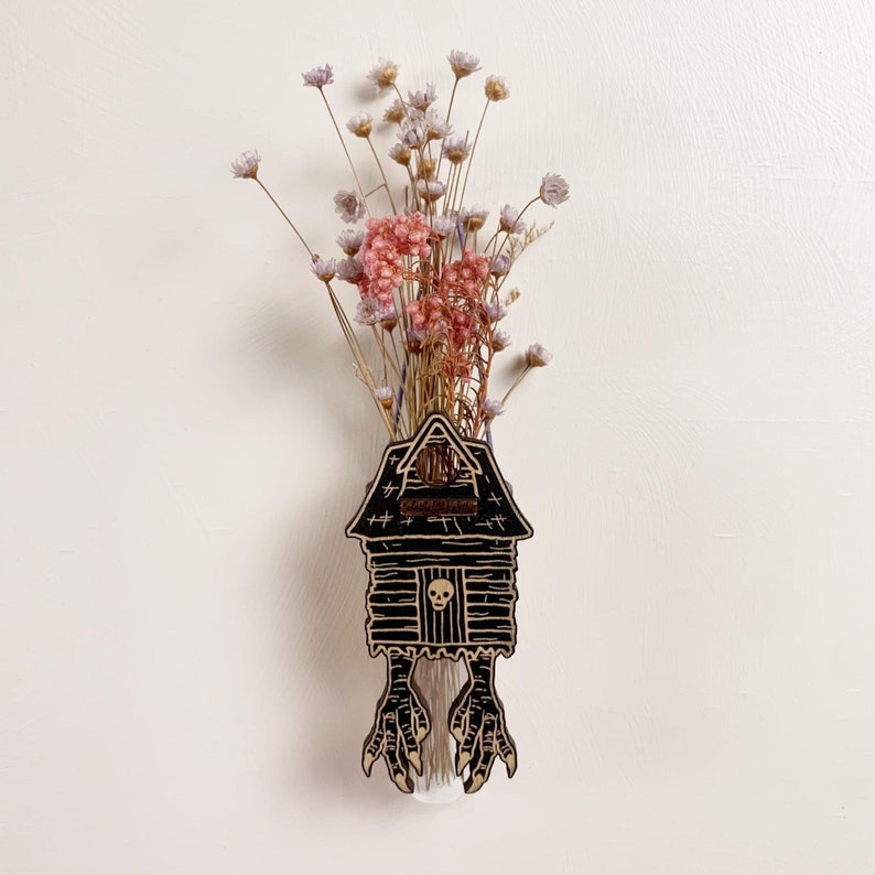 May include: A unique wooden house-shaped vase with a black finish, featuring a skull and claw feet design. The vase holds dried flowers in shades of pink and purple. A creative and spooky home decor item.