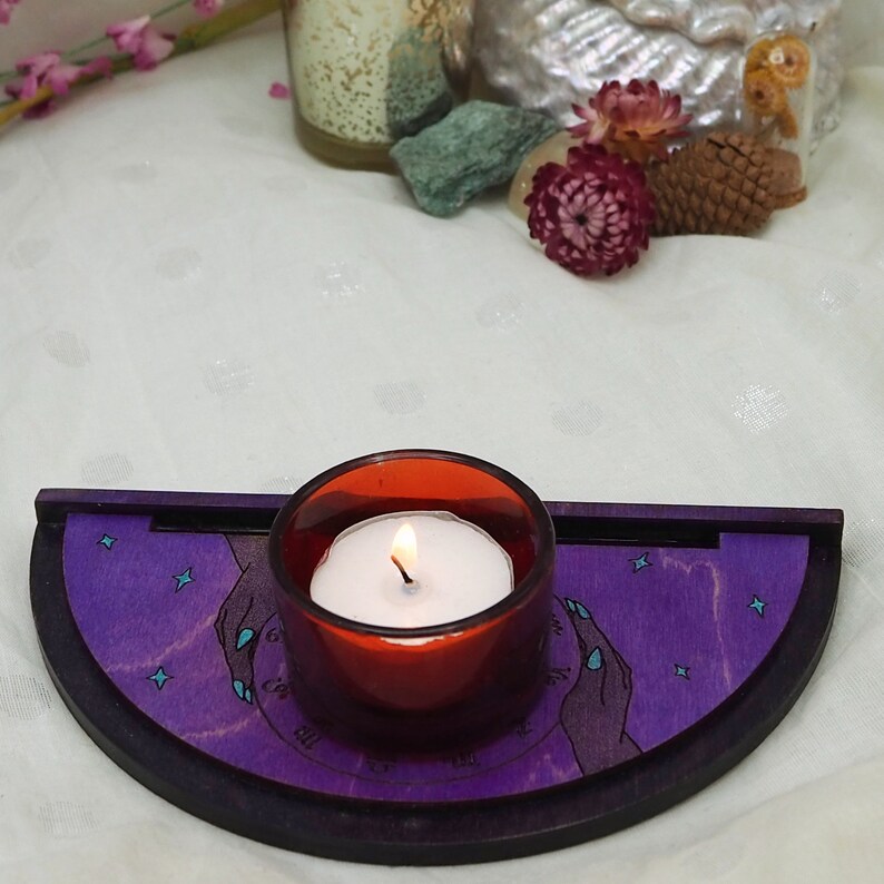 Puede incluir: Un portavelas de madera morado con forma de media luna y un peque&ntilde;o portavelas de vidrio rojo con una vela blanca dentro. El portavelas tiene un dise&ntilde;o de dos manos con estrellas en el fondo.