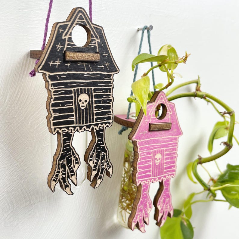May include: Two wooden house-shaped planters, one black and one pink, each with a skull and bird feet design. The planters hang from twine and hold glass vases with plants. The houses have a rustic, spooky aesthetic.